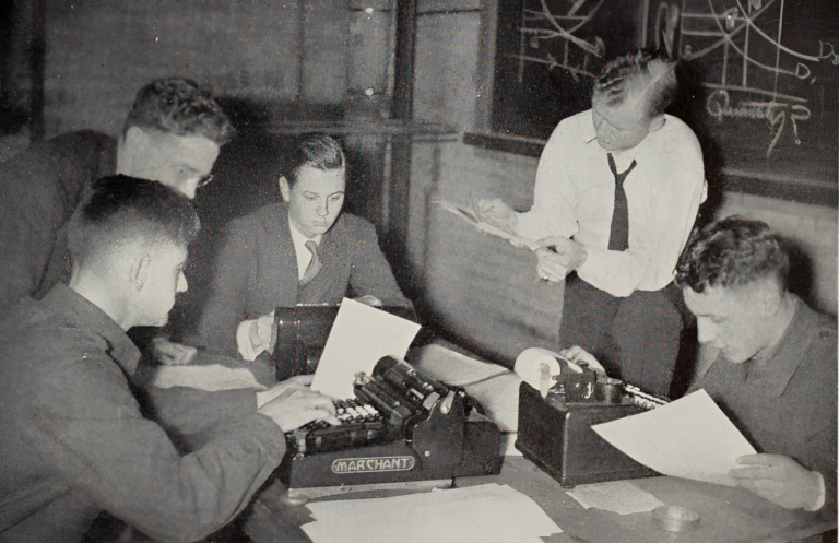 Black and white photo of five students grouped around a table using large old-fashioned calculators.