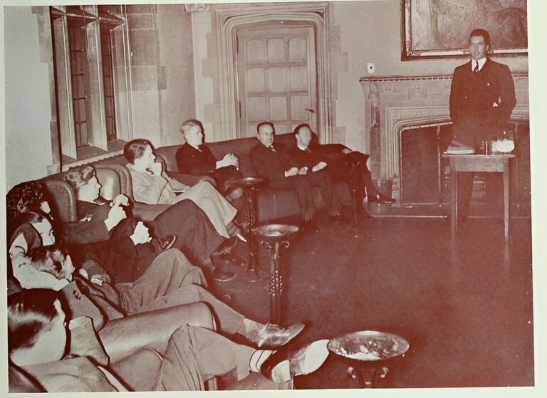 Black and white photo of group of people sitting on couches with one person standing by small table.