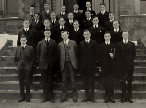 Black and white photo of a group of men in suits standing by steps outside of a building.