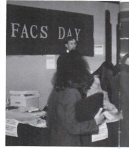 Black and white photo of person talking to a group of students with sign behind them that reads FACS Day.