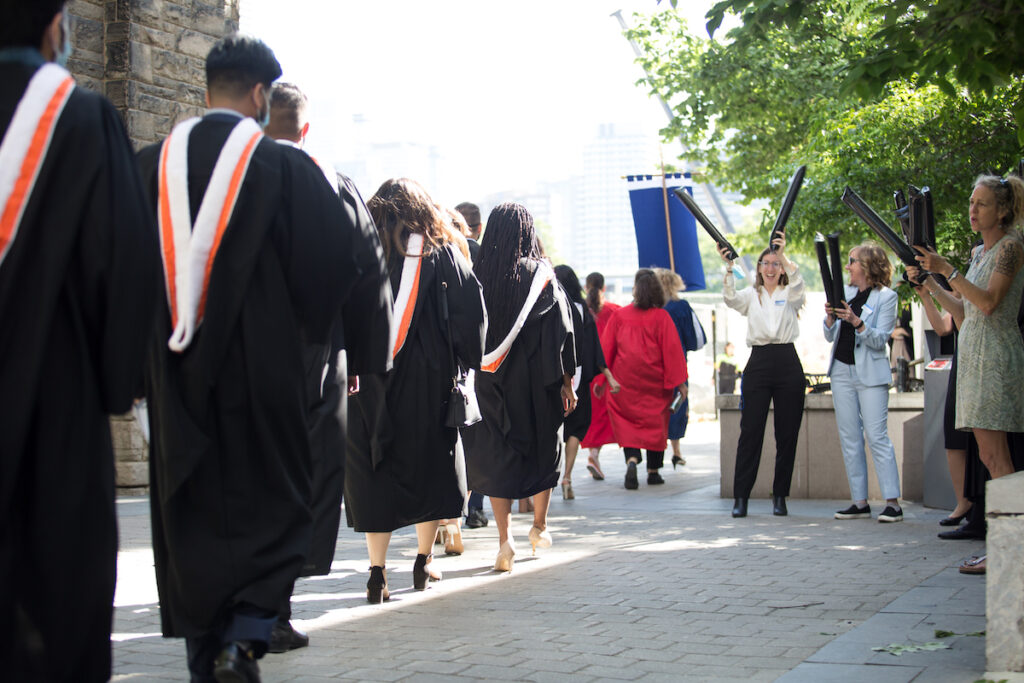 Students dressed in graduation gowns walking in a line with a group of people on the right cheering them on with black inflatable noisemakers.