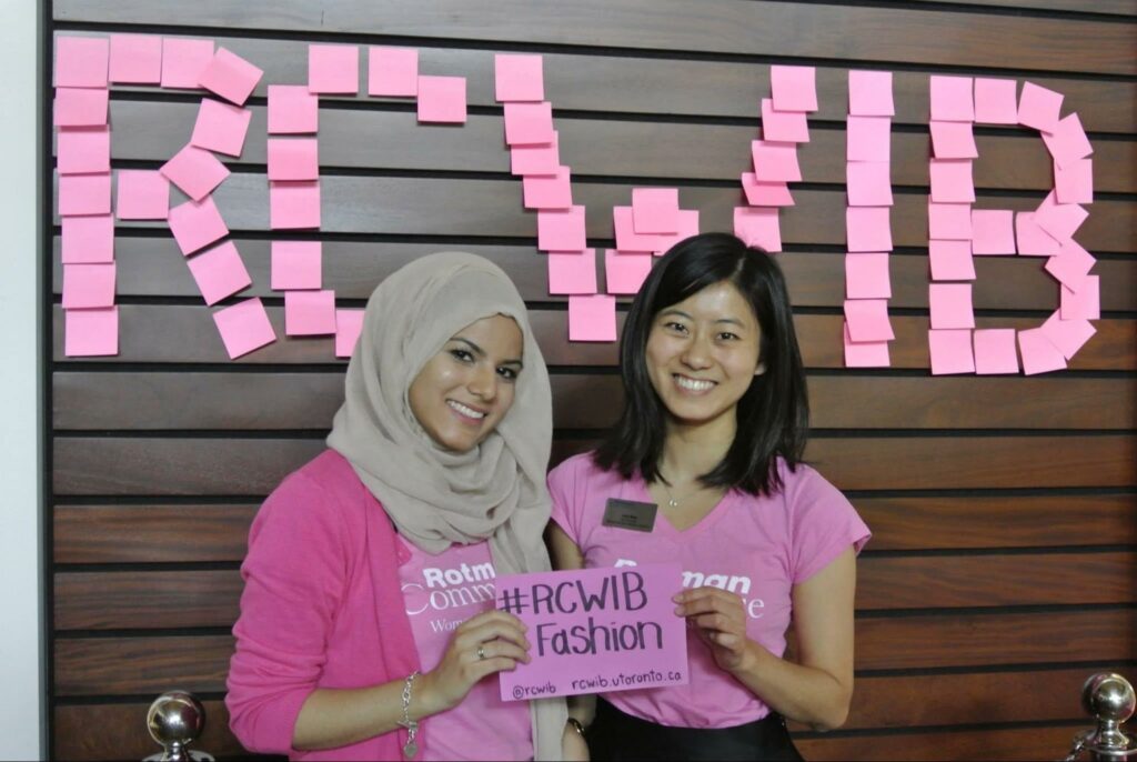 Two people wearing pink Rotman Commerce shirts and holding a paper between them that says ‘#RCWIB Fashion’.