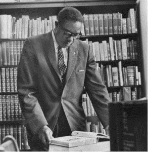 Black and white photo of man in a suit standing at a desk reading a book with a book shelf with books in the background.