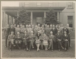 Black and white photo with large group of people with the front row sitting in chairs and the two back rows standing. They are standing in front of a building with a front porch.